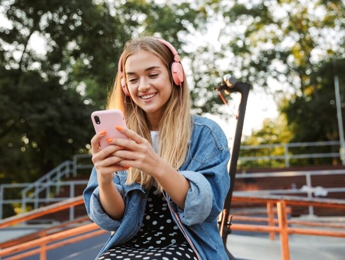A young teen looks down at her phone smiling with headphones on at a park.