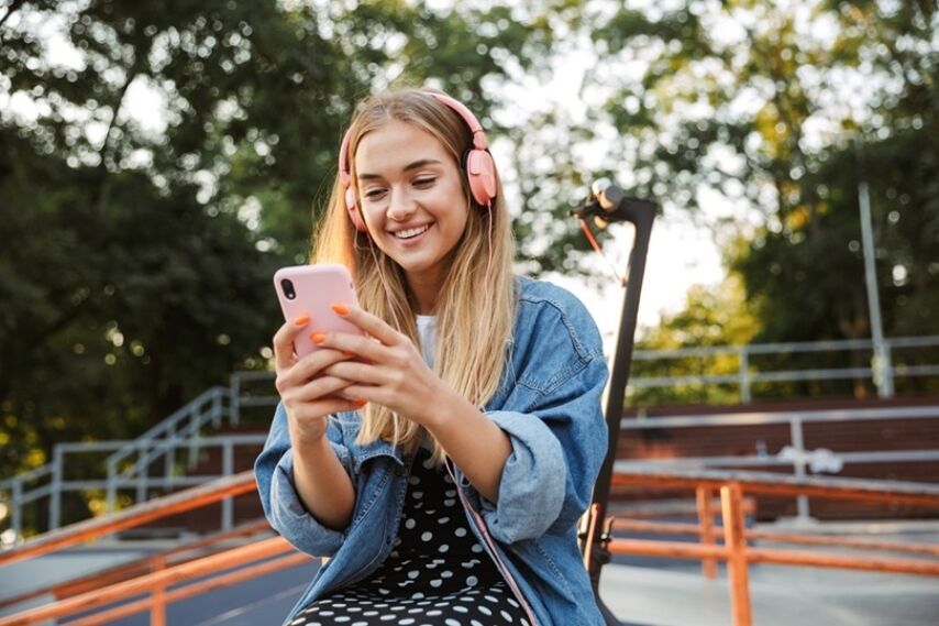 A young teen looks down at her phone smiling with headphones on at a park.