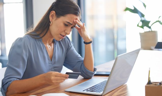 A stressed woman looks at her credit card with her laptop open.