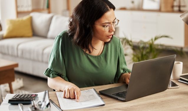 A woman works on a laptop while referencing a notebook