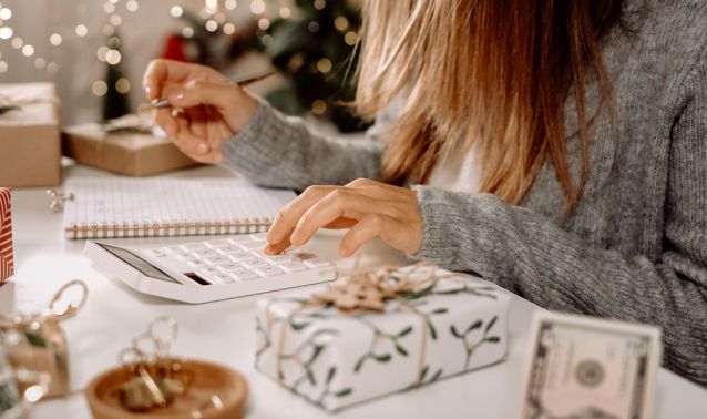 Hands use a calculator and pen at a table surrounded by wrapped gifts, a notebook, and currency, suggesting someone planning or budgeting for holiday expenses in a warm, festive home setting.