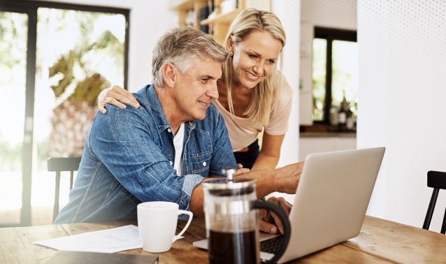 Two adults sit closely at a wooden table in a bright, modern kitchen, looking at a laptop together. Papers, a coffee mug, and a French press are on the table, suggesting they are reviewing or working on something collaboratively.