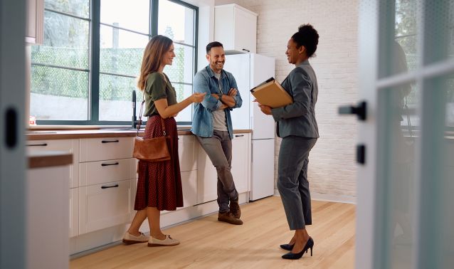 Three adults stand and talk in a modern, well-lit kitchen with white cabinets and large windows. One person holds a clipboard while the others listen and gesture, suggesting a discussion or consultation about the space.