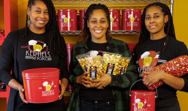 Three people stand indoors holding branded popcorn products, including red popcorn tins and clear bags of flavored popcorn, with shelves of matching red containers displayed behind them against a bright yellow wall.