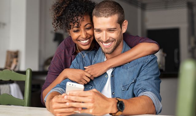 A couple sits closely at a table in a cozy home setting, with one person hugging the other from behind while they look at a smartphone together, creating a warm, relaxed moment.