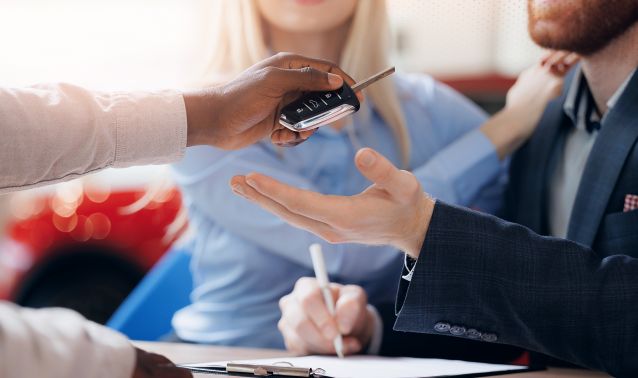 A person hands car keys to another person across a desk while paperwork is being signed, with a couple seated together, suggesting the completion of a car purchase or financing agreement.