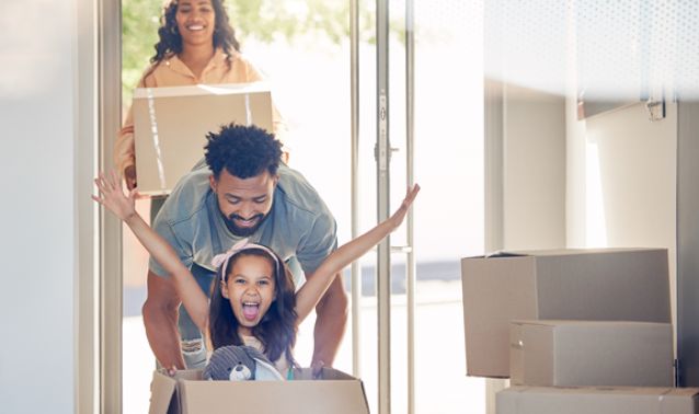 A family moves into a new home, with two adults carrying boxes through a sunlit doorway while a child sits inside a cardboard box with arms raised playfully, surrounded by stacked moving boxes.