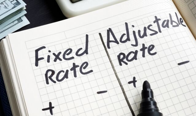 A notebook on a desk shows a handwritten comparison between “Fixed Rate” and “Adjustable Rate,” with plus and minus symbols under each heading. A black marker rests on the page, and money and a calculator are partially visible nearby, suggesting a financial or mortgage-related decision.