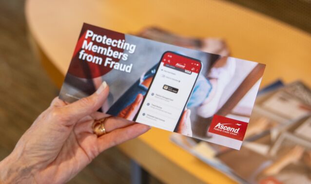 A hand holds a brochure titled “Protecting Members from Fraud,” featuring a smartphone displaying the Ascend Federal Credit Union mobile app. The background shows a table with magazines.