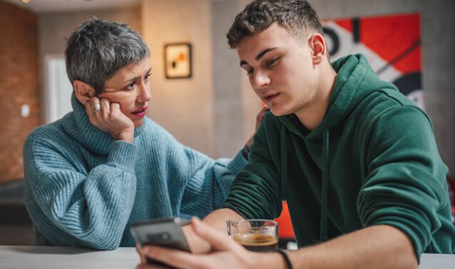 Two people sit at a table indoors, one holding a smartphone and a glass of coffee. The other person rests a hand near their face while leaning closer, appearing to look at the phone. A modern living space with artwork is visible in the background.