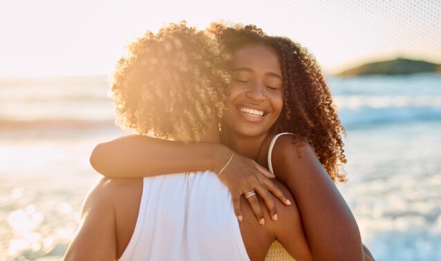 Two adults embracing near an ocean shoreline at sunset, photographed from behind one person, with waves and warm sunlight in the background.