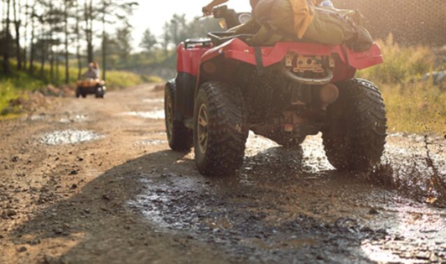 Two all-terrain vehicles driving along a muddy dirt road in a wooded area, with water splashing from the tires and one vehicle visible farther ahead.