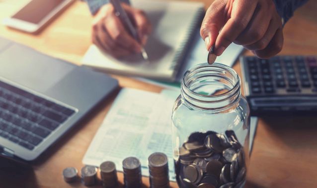 Hands putting change into a Mason jar while using a calculator and laptop.