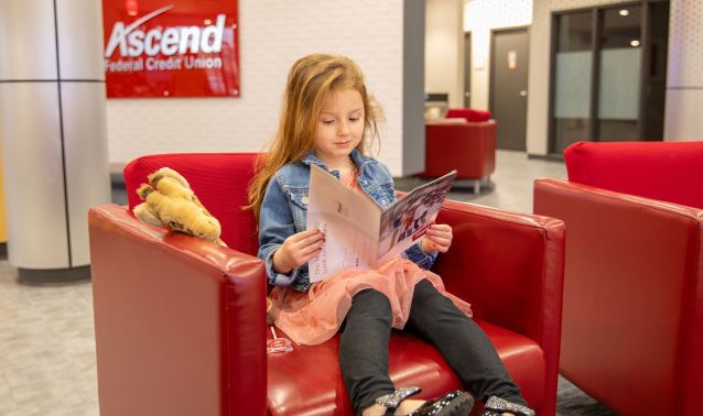 A young child sits at a low table in a modern lobby, coloring in an activity book while reaching out to touch a plush giraffe toy. Red lounge chairs and an Ascend sign in the background create a welcoming, family-friendly environment.