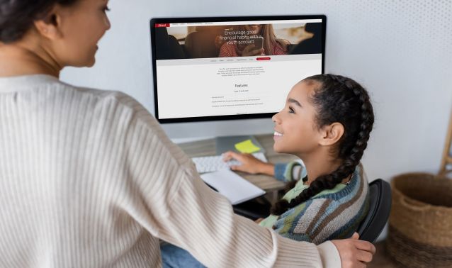 An adult stands beside a child seated at a desk, with a supportive hand on the child’s shoulder, as they look at a desktop computer displaying a financial services webpage. Sticky notes and office items surround the workspace, suggesting a guided learning or assistance moment at home.
