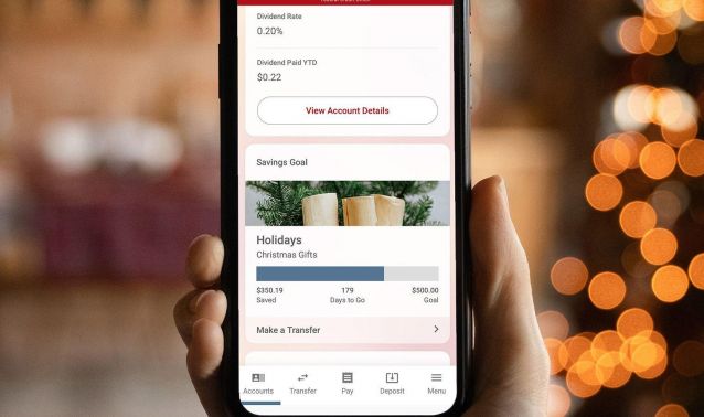 A hand holds a smartphone displaying the Ascend mobile banking app, showing account details, a savings goal labeled “Holidays,” and options to view account details or make a transfer, with warm, out-of-focus lights in the background.