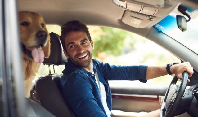 A man smiles in his car with his golden retriever beside him.