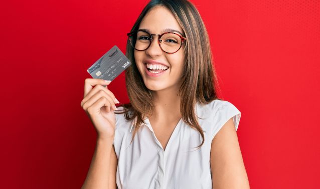 A person stands against a bold red background holding a credit card near their shoulder, wearing a white short-sleeve blouse. The image has a clean, modern look that emphasizes personal finance or card-based banking services.