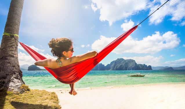 A person relaxes in a red hammock tied between a palm tree and a post on a sandy beach, overlooking clear turquoise water and distant rocky islands. Bright sunlight and scattered clouds create a peaceful tropical scene, with a small boat visible on the horizon.