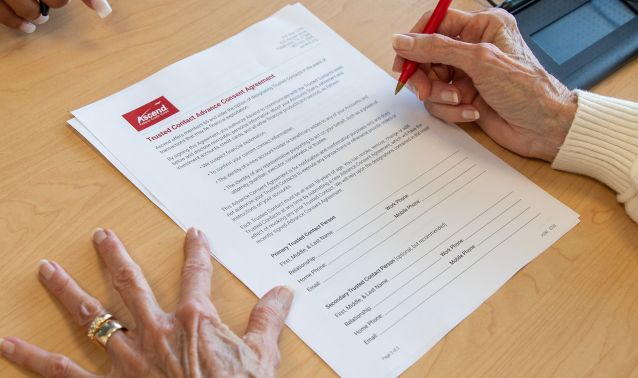 Hands rest on a wooden table as one person uses a red pen to review and sign a printed financial agreement document with an Ascend logo at the top, while another person gestures nearby, suggesting a guided discussion or assistance with paperwork.