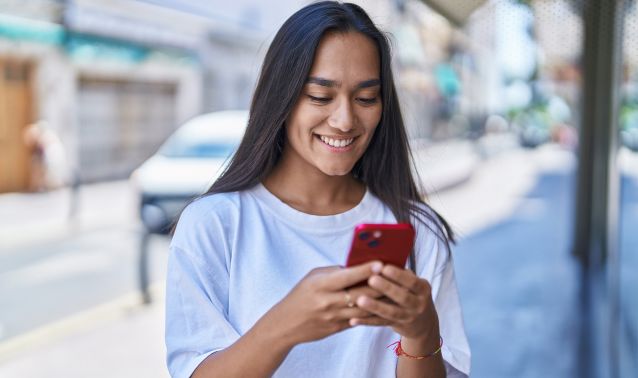 A person stands on a city sidewalk holding and looking at a red smartphone, wearing a white T‑shirt. Parked cars, storefronts, and a covered walkway line the street in the background, suggesting an everyday urban setting.