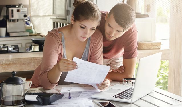 Two people sit together at a kitchen table reviewing paperwork, with one holding a document while the other leans in to read along. A laptop, smartphone, coffee maker, and scattered papers are on the table, suggesting they are organizing finances or going over household bills in a home setting.
