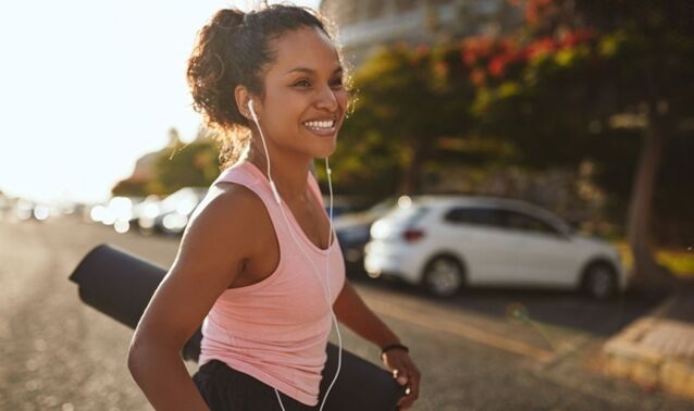 A woman walks across a parking lot smiling with earbuds in and a bag over her shoulder.