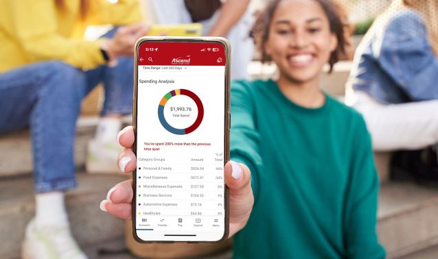 A person holds a smartphone toward the camera displaying the Ascend mobile app’s spending analysis screen, with a colorful chart and total spending amount visible. Other people sit casually in the background, creating a relaxed, social setting while highlighting the app’s financial insights.
