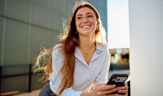 A woman looks off in the distance smiling with a phone in her hand.