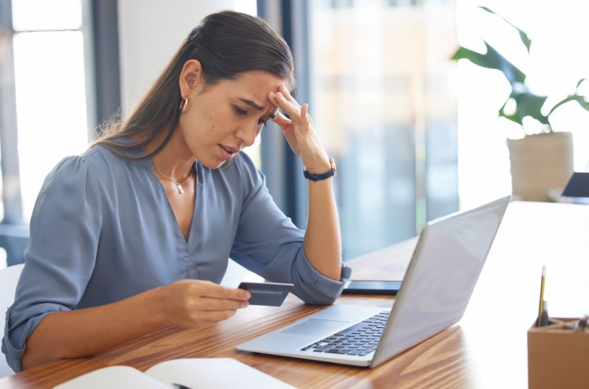 A stressed woman looks at her credit card with her laptop open.