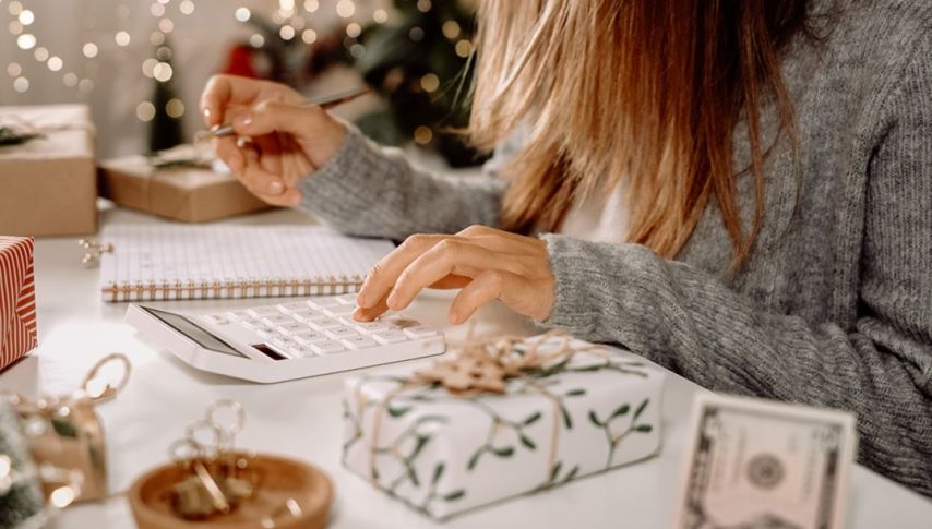 Hands use a calculator and pen at a table surrounded by wrapped gifts, a notebook, and currency, suggesting someone planning or budgeting for holiday expenses in a warm, festive home setting.