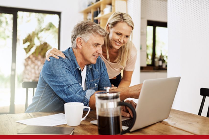Two adults sit closely at a wooden table in a bright, modern kitchen, looking at a laptop together. Papers, a coffee mug, and a French press are on the table, suggesting they are reviewing or working on something collaboratively.