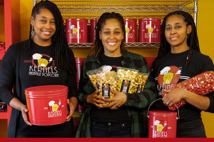 Three people stand indoors holding branded popcorn products, including red popcorn tins and clear bags of flavored popcorn, with shelves of matching red containers displayed behind them against a bright yellow wall.