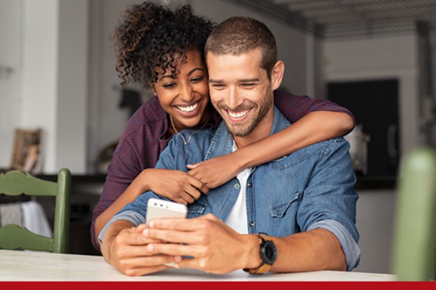 A couple sits closely at a table in a cozy home setting, with one person hugging the other from behind while they look at a smartphone together, creating a warm, relaxed moment.