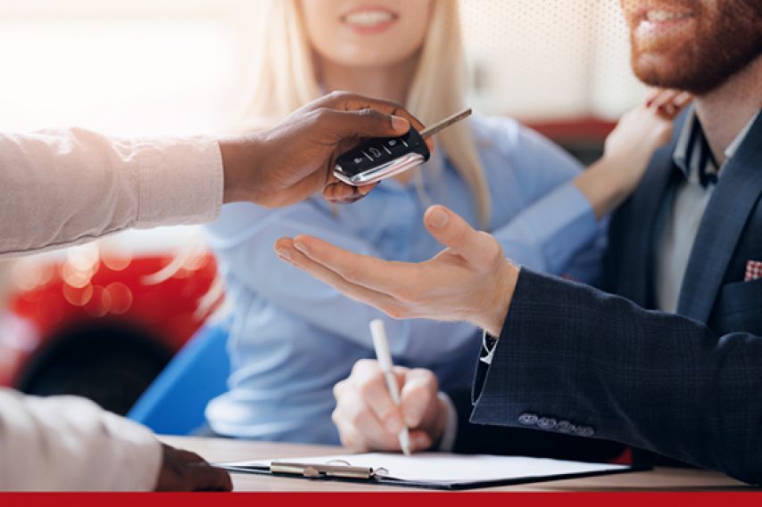 A person hands car keys to another person across a desk while paperwork is being signed, with a couple seated together, suggesting the completion of a car purchase or financing agreement.