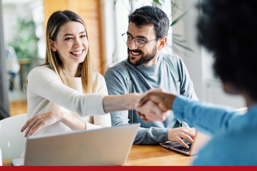 Two people sit at a table with laptops during a meeting, reaching across the table to shake hands with a third person, suggesting a successful agreement or professional discussion in a bright, modern workspace.