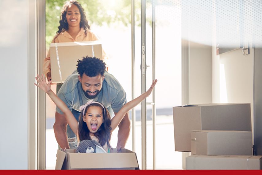 A family moves into a new home, with two adults carrying boxes through a sunlit doorway while a child sits inside a cardboard box with arms raised playfully, surrounded by stacked moving boxes.