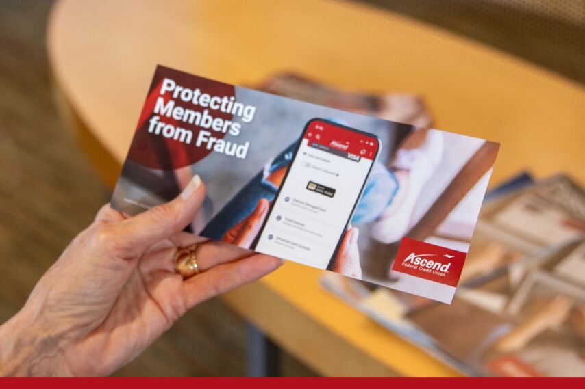 A hand holds a brochure titled “Protecting Members from Fraud,” featuring a smartphone displaying the Ascend Federal Credit Union mobile app. The background shows a table with magazines.