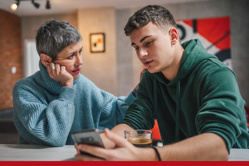 Two people sit at a table indoors, one holding a smartphone and a glass of coffee. The other person rests a hand near their face while leaning closer, appearing to look at the phone. A modern living space with artwork is visible in the background.
