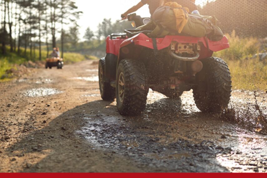 Two all-terrain vehicles driving along a muddy dirt road in a wooded area, with water splashing from the tires and one vehicle visible farther ahead.