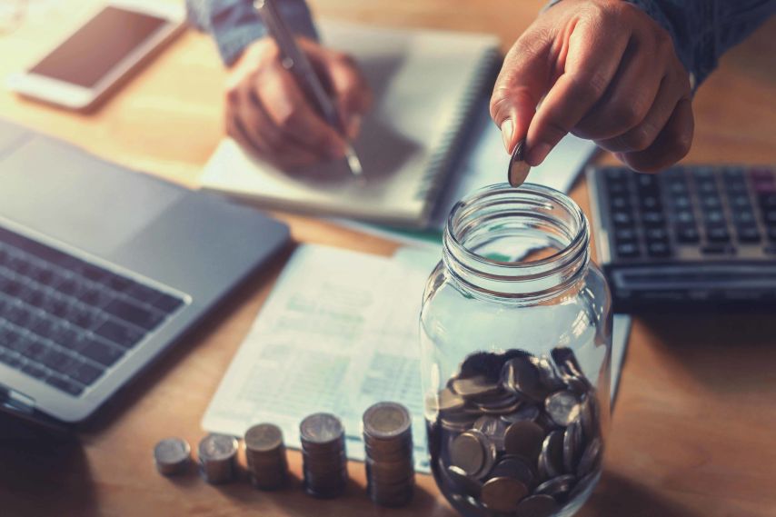 Hands putting change into a Mason jar while using a calculator and laptop.