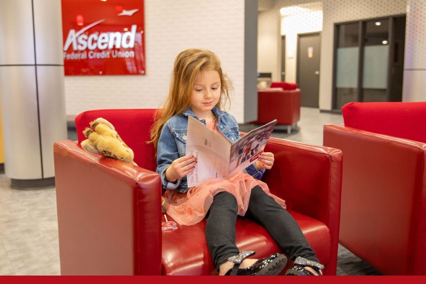 A young child sits at a low table in a modern lobby, coloring in an activity book while reaching out to touch a plush giraffe toy. Red lounge chairs and an Ascend sign in the background create a welcoming, family-friendly environment.