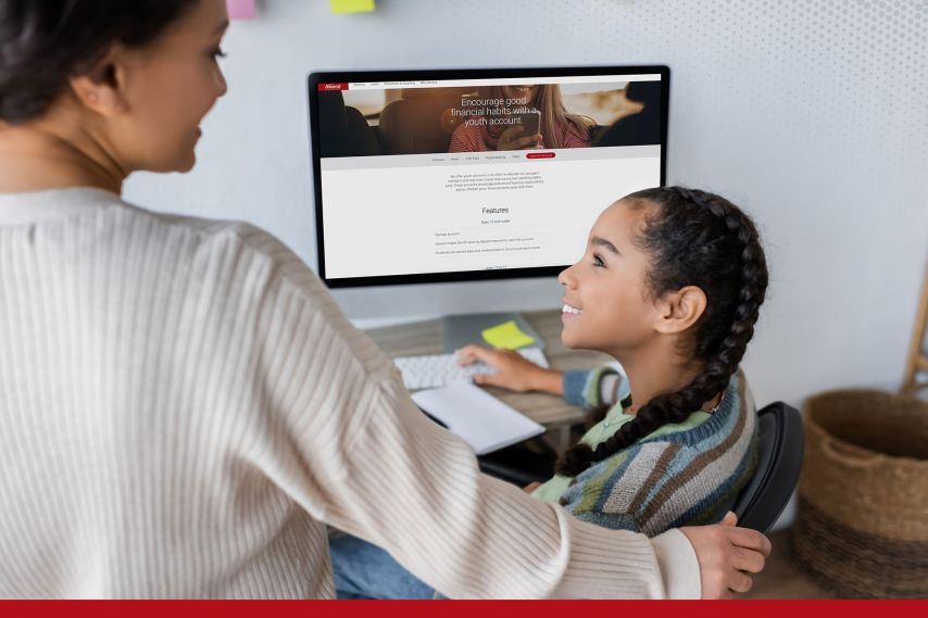 An adult stands beside a child seated at a desk, with a supportive hand on the child’s shoulder, as they look at a desktop computer displaying a financial services webpage. Sticky notes and office items surround the workspace, suggesting a guided learning or assistance moment at home.