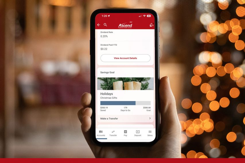 A hand holds a smartphone displaying the Ascend mobile banking app, showing account details, a savings goal labeled “Holidays,” and options to view account details or make a transfer, with warm, out-of-focus lights in the background.