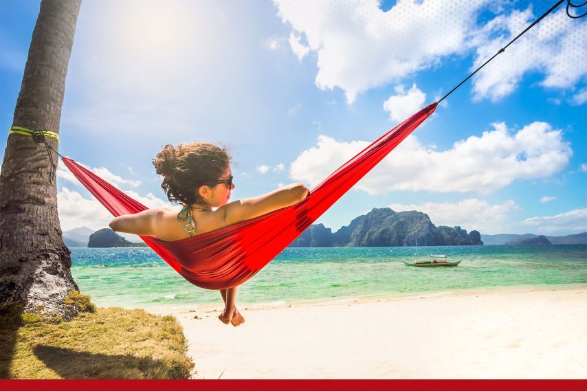 A person relaxes in a red hammock tied between a palm tree and a post on a sandy beach, overlooking clear turquoise water and distant rocky islands. Bright sunlight and scattered clouds create a peaceful tropical scene, with a small boat visible on the horizon.