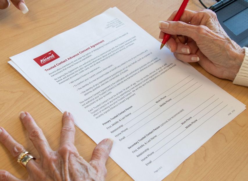 Hands rest on a wooden table as one person uses a red pen to review and sign a printed financial agreement document with an Ascend logo at the top, while another person gestures nearby, suggesting a guided discussion or assistance with paperwork.