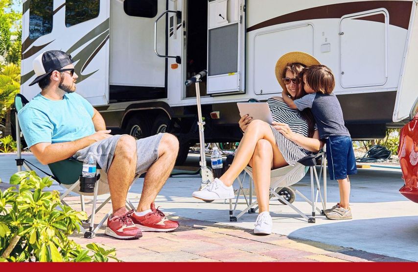 A family relaxes at a campsite beside a large RV, with adults seated in folding chairs and a child standing close while looking at a tablet together. A red car is parked nearby, and the sunny outdoor setting suggests a casual, leisurely moment during a road trip or camping stay