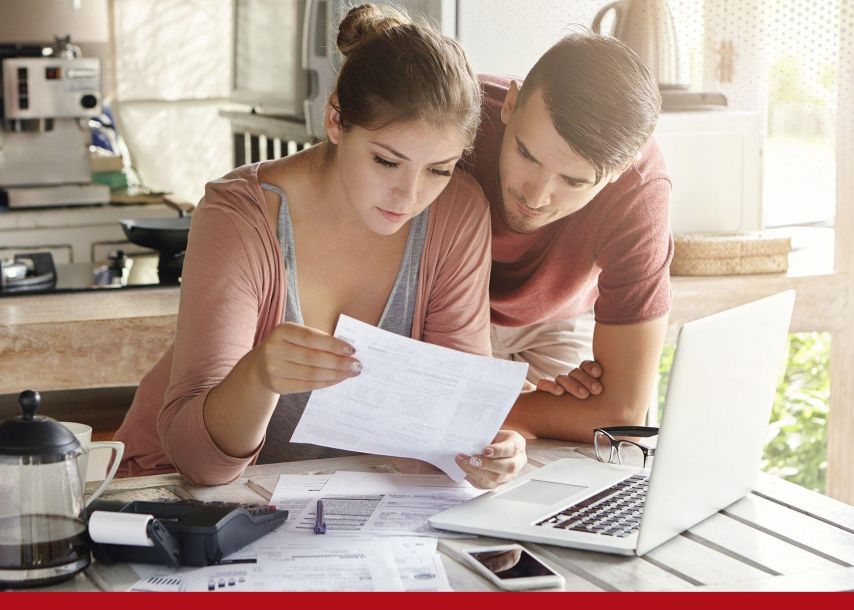 Two people sit together at a kitchen table reviewing paperwork, with one holding a document while the other leans in to read along. A laptop, smartphone, coffee maker, and scattered papers are on the table, suggesting they are organizing finances or going over household bills in a home setting.