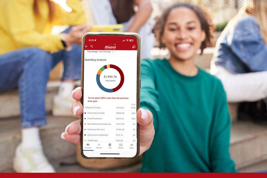 A person holds a smartphone toward the camera displaying the Ascend mobile app’s spending analysis screen, with a colorful chart and total spending amount visible. Other people sit casually in the background, creating a relaxed, social setting while highlighting the app’s financial insights.